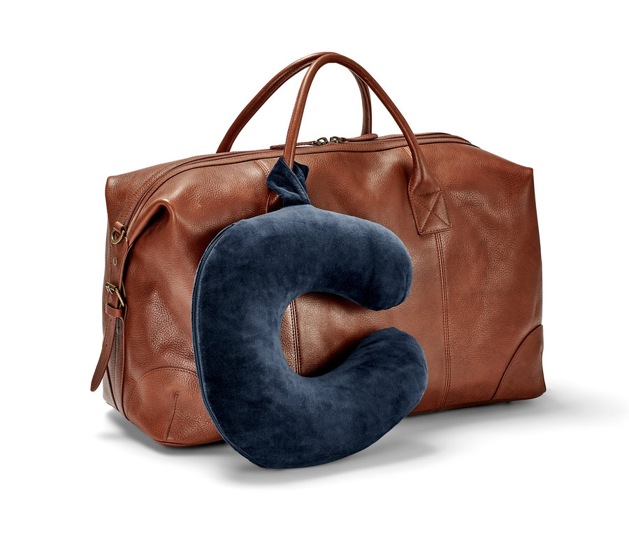Ein dunkelblaues Nackenkissen aus Memory Foam lehnt an einer braunen Reisetasche aus Leder.