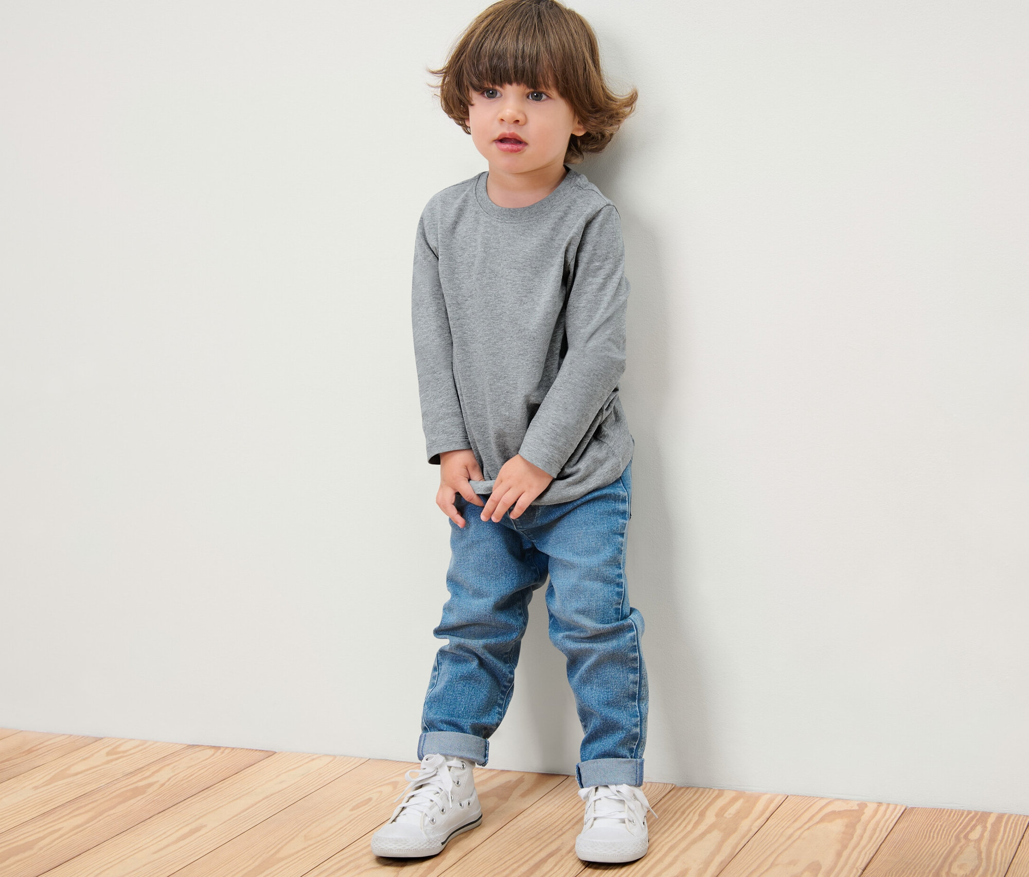 Ein Junge in einem grauen Langarmshirt (5 Kinder-Langarmshirts), Jeans und weißen Schuhen steht an einer Wand.