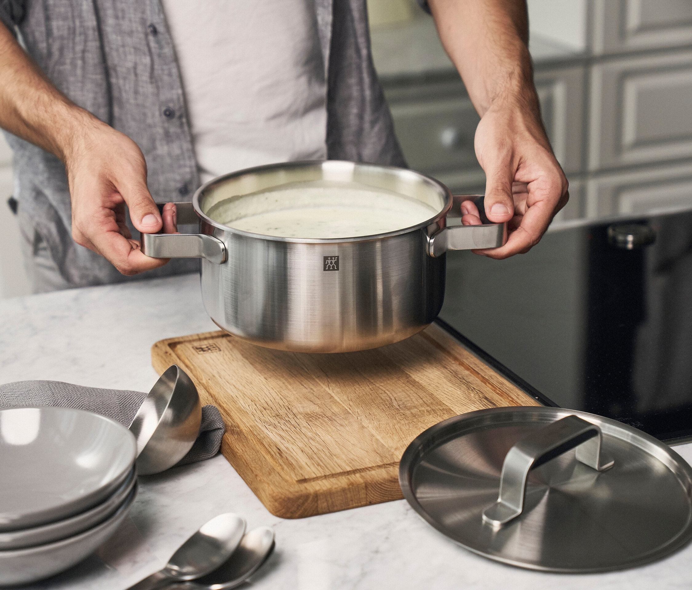 Mann hält einen silberfarbenen ZWILLING PURE Kochtopf aus einem 5-teiligen Kochgeschirr-Set aus Edelstahl über einem Holzbrett. Graue Schüsseln, eine Schöpfkelle, Löffel, Gläser und ein Deckel stehen daneben.