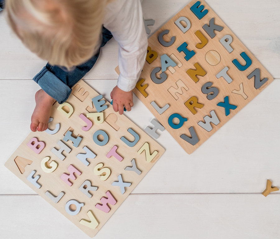 Ein Kind spielt mit zwei hölzernen ABC-Puzzles »Hanna« und ABC-Puzzles »Caspar« auf einer weißen Holzoberfläche.