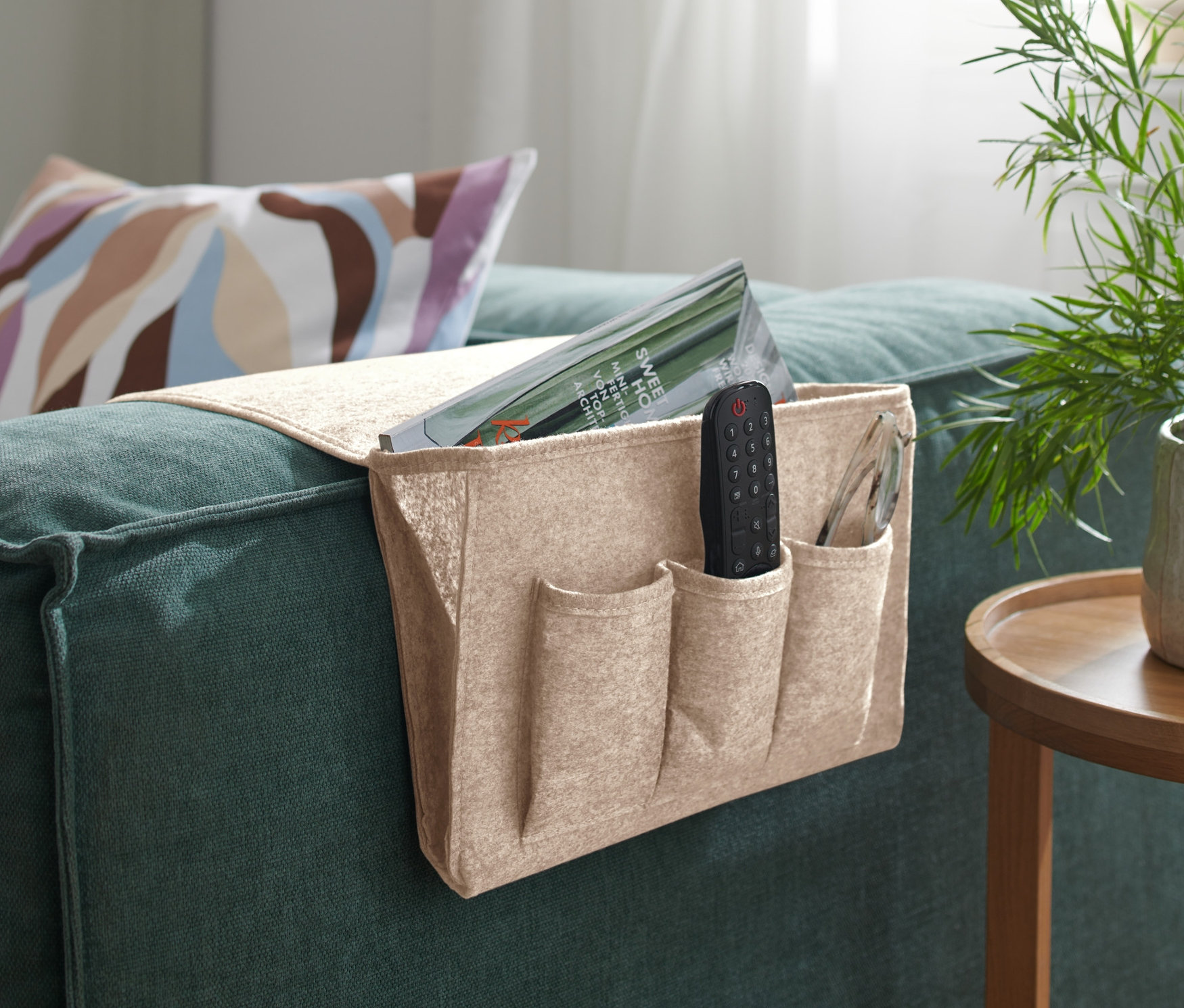 Ein beiger Bett- und Couchorganizer hängt an der Armlehne eines grünen Sofas, darin befinden sich eine Fernbedienung und eine Zeitschrift.