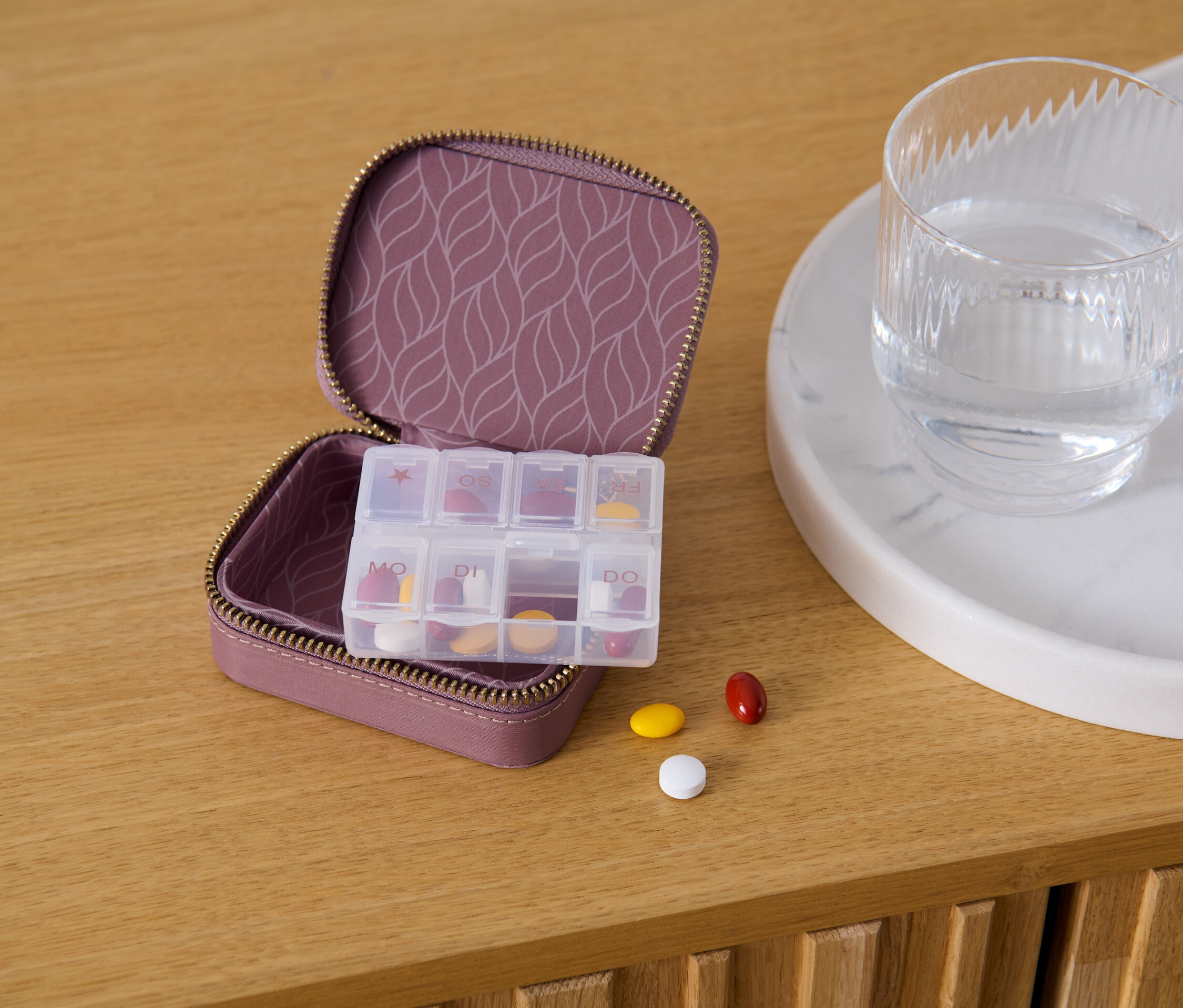 Offene lilafarbene Pillendose mit gemustertem Innenraum, daneben ein Glas Wasser und drei Pillen auf einem Holztisch.