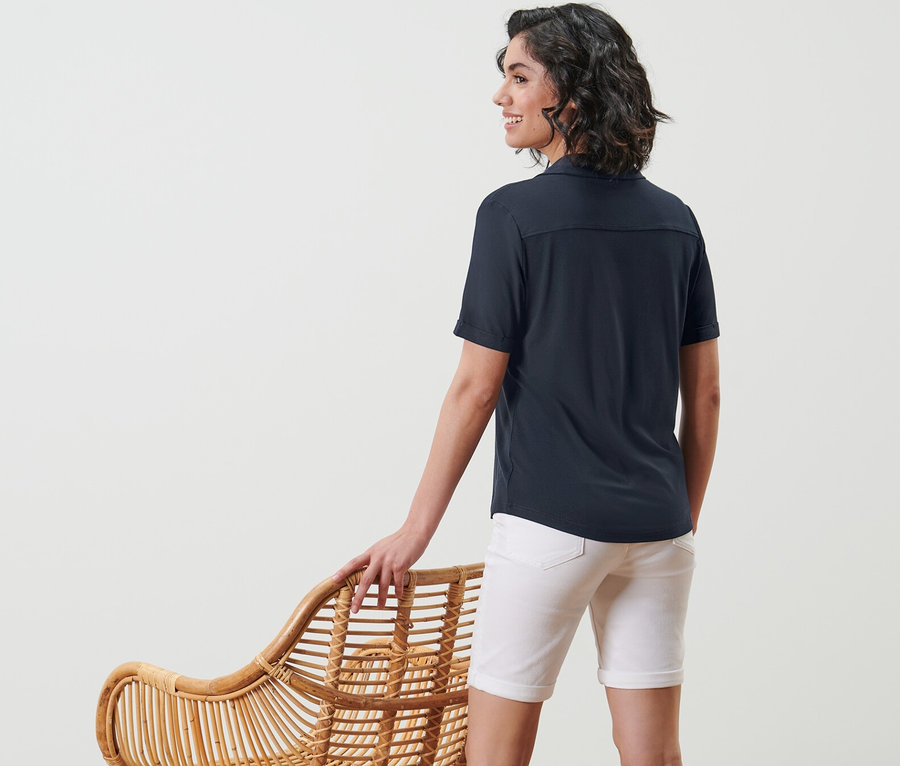 Eine Frau steht an einem Korbsessel und trägt ein dunkelblaues Blusenshirt mit Knopfleiste.