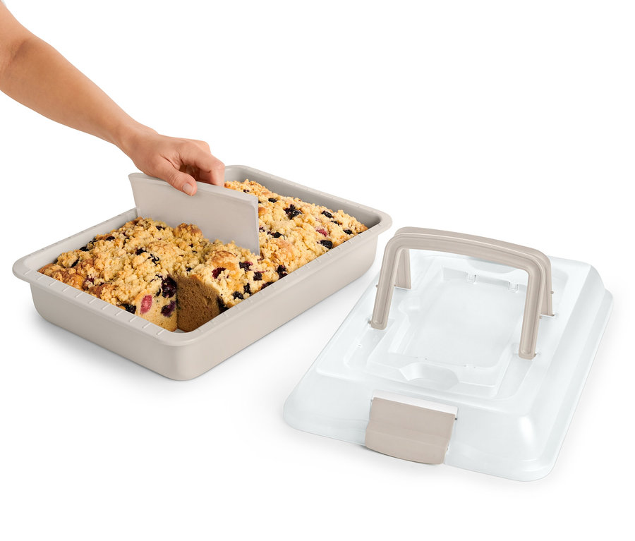 Eine Hand schneidet einen Obstkuchen in einem beigen Backblech mit Transportdeckel.