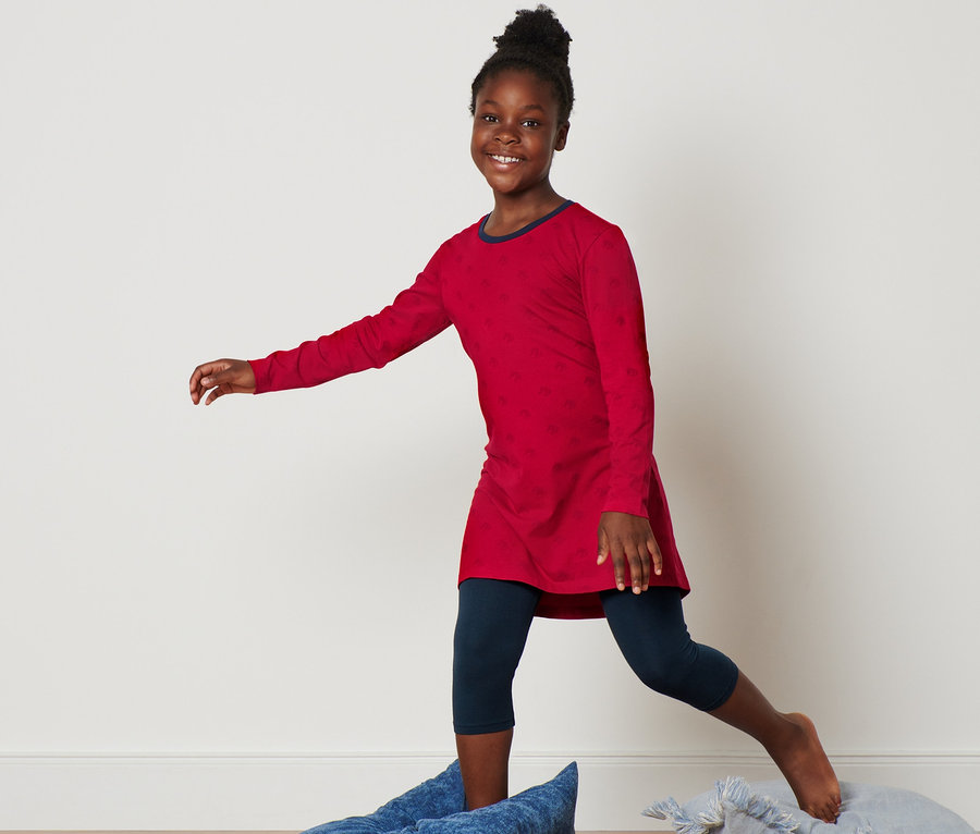 Ein lächelndes Mädchen balanciert auf Kissen. Sie trägt ein rotes Nachthemd und eine 3/4-Leggings.