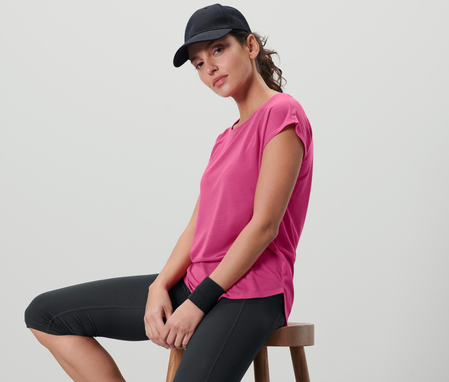 Frau sitzt auf einem Hocker und trägt ein fuchsiafarbenes Sportshirt, schwarze Sportleggings und eine schwarze Baseballkappe.
