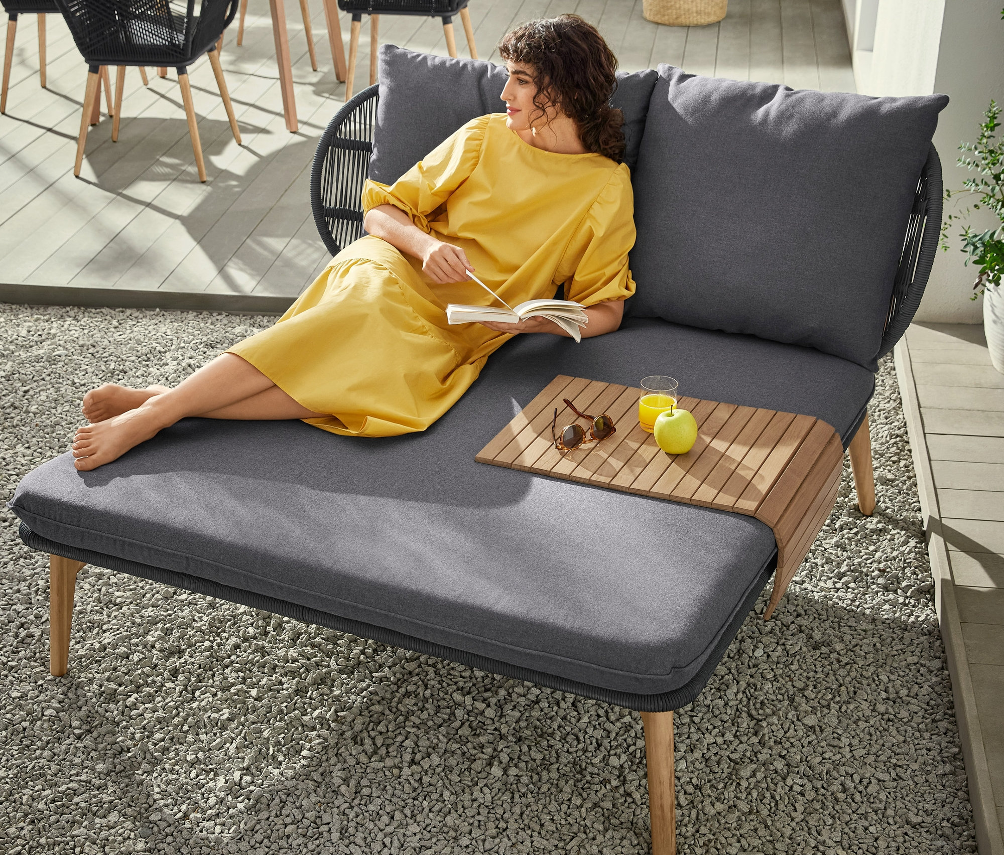 Eine Frau im gelben Kleid liegt auf einem Daybed mit Kopfteil mit Textilgeflecht und liest ein Buch. Auf dem Daybed stehen ein Tischchen, ein Getränk und ein Apfel. Im Hintergrund stehen ein runder Tisch, ein Schalenstuhl mit Textilgeflecht und eine LED-Akku-Tischleuchte.