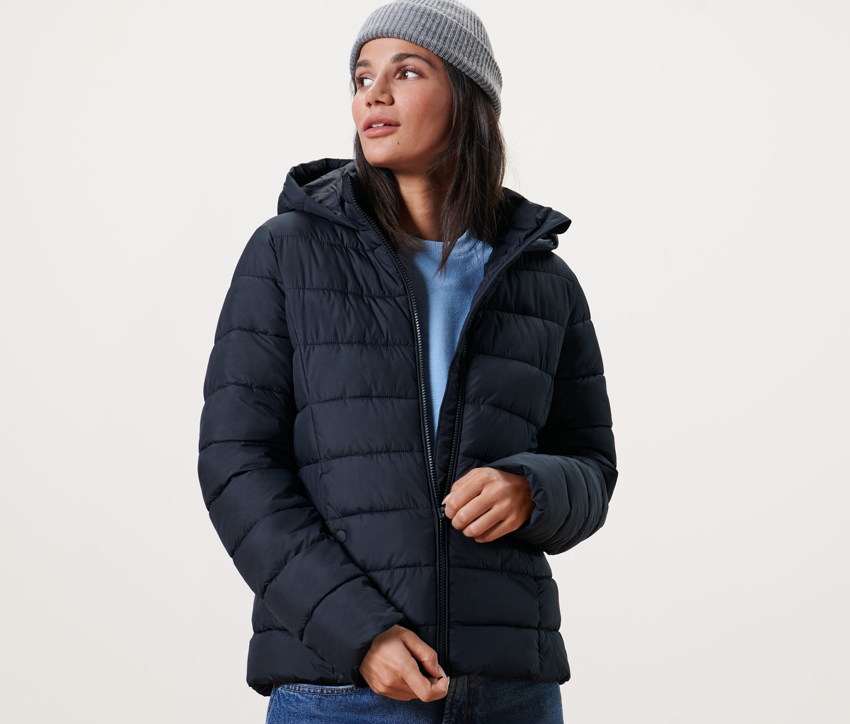 Frau schliesst eine dunkelblaue Steppjacke mit Kapuze und trägt eine graue Strickmütze.