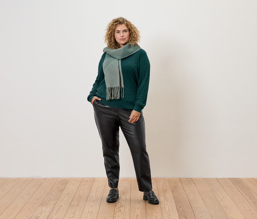 Frau mit lockigem Haar posiert in einem grünen Pullover, Schal und schwarzen Lederhosen.