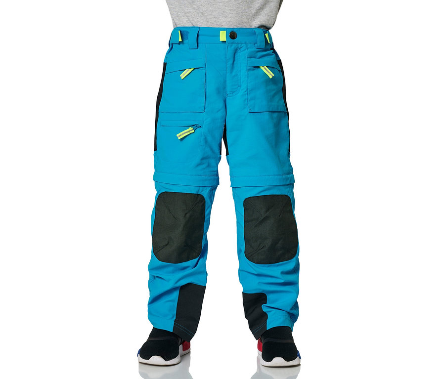 Ein Junge trägt eine blaue Jako-O Zipp-off-Hose mit Taschen und gelben Details, er steht vor einem weißen Hintergrund.