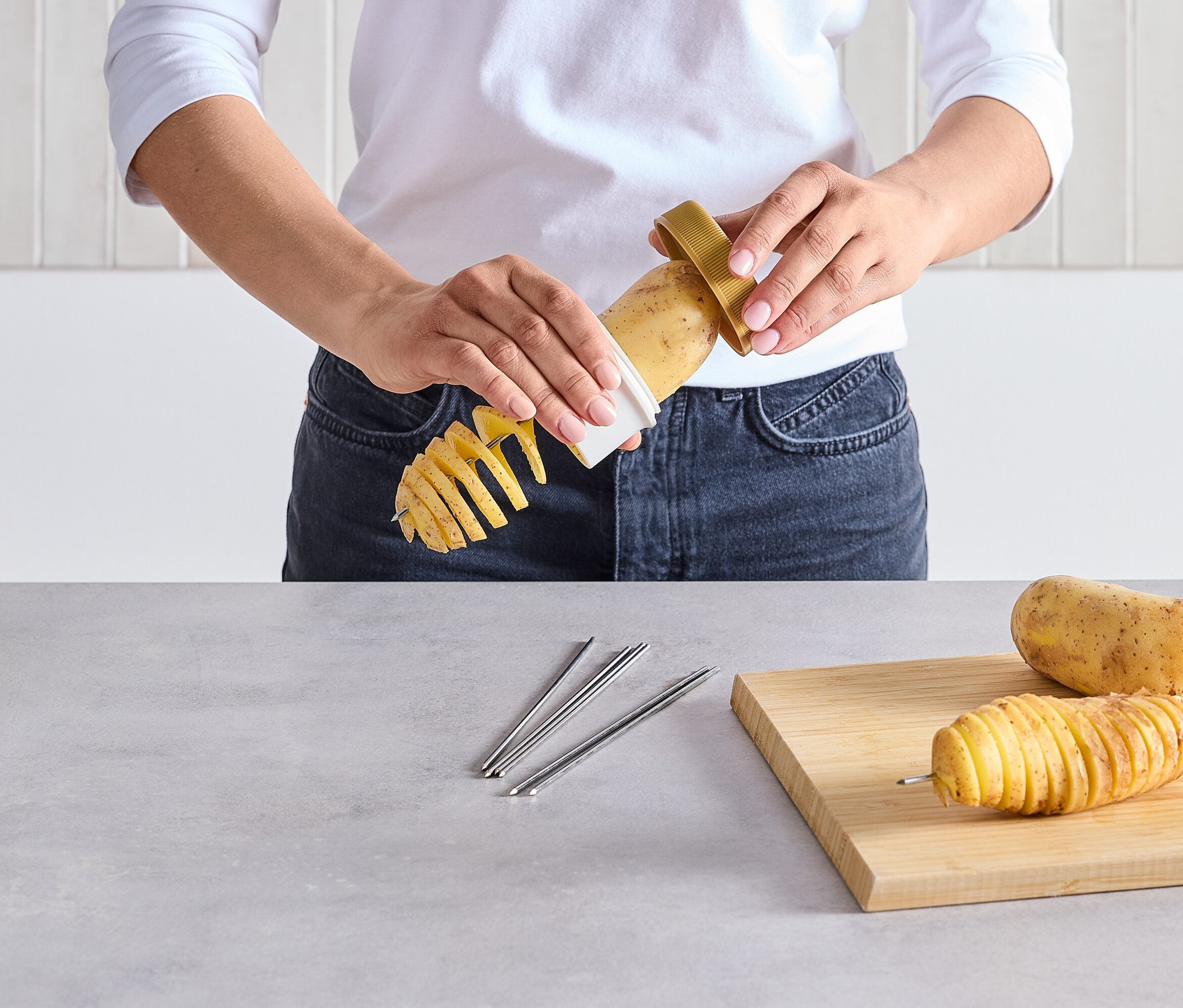 Eine Person schneidet eine Kartoffel mit einem Spiralschneider. Auf dem Tisch liegen eine geschnittene Kartoffel am Spieß und eine rohe Kartoffel.