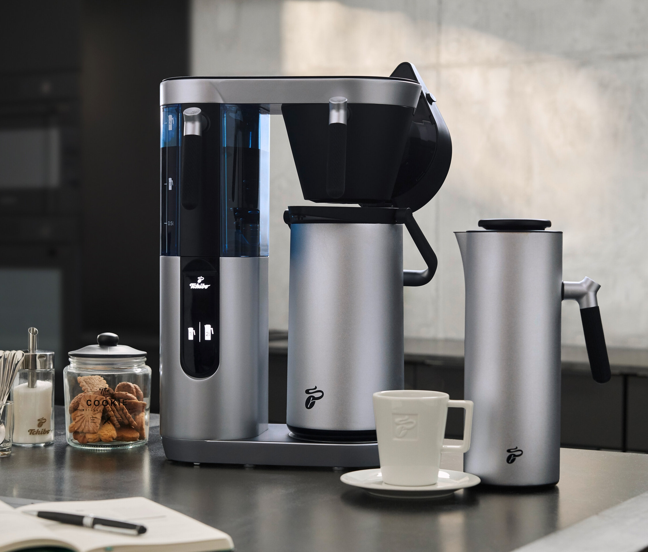 Silberne Profi-Filterkaffeemaschine Coffea Filbii neben Filbii 1L und 2L Thermoskannen. Auf dem Tisch liegen ein offenes Notizbuch und Gläser mit Zucker und Keksen.