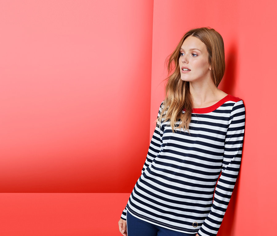 Frau lehnt an roter Wand, trägt gestreiftes Langarmshirt und blaue Hose.