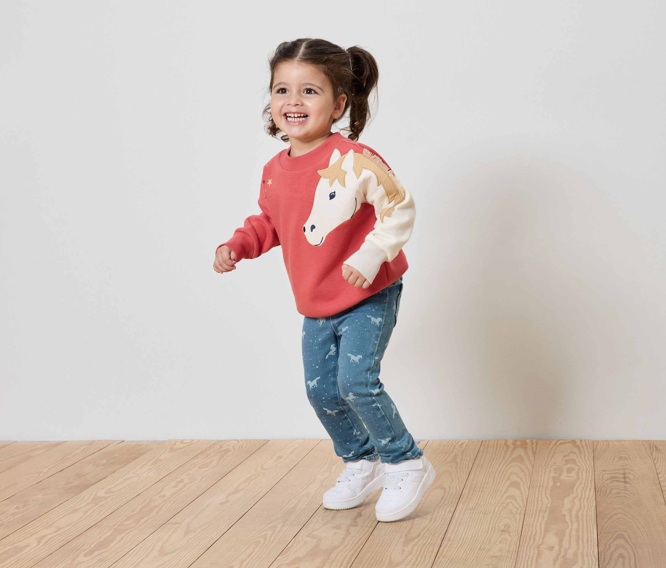 Lächelndes kleines Mädchen in einem korallenroten Pullover mit Pferdemotiv und blauen Jeans mit Aufdruck steht auf einem Holzboden.