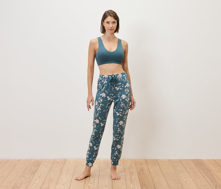 Frau steht barfuß vor einer Wand und trägt einen blauen Sport-BH und eine blaue Jogginghose mit Blumenmuster.