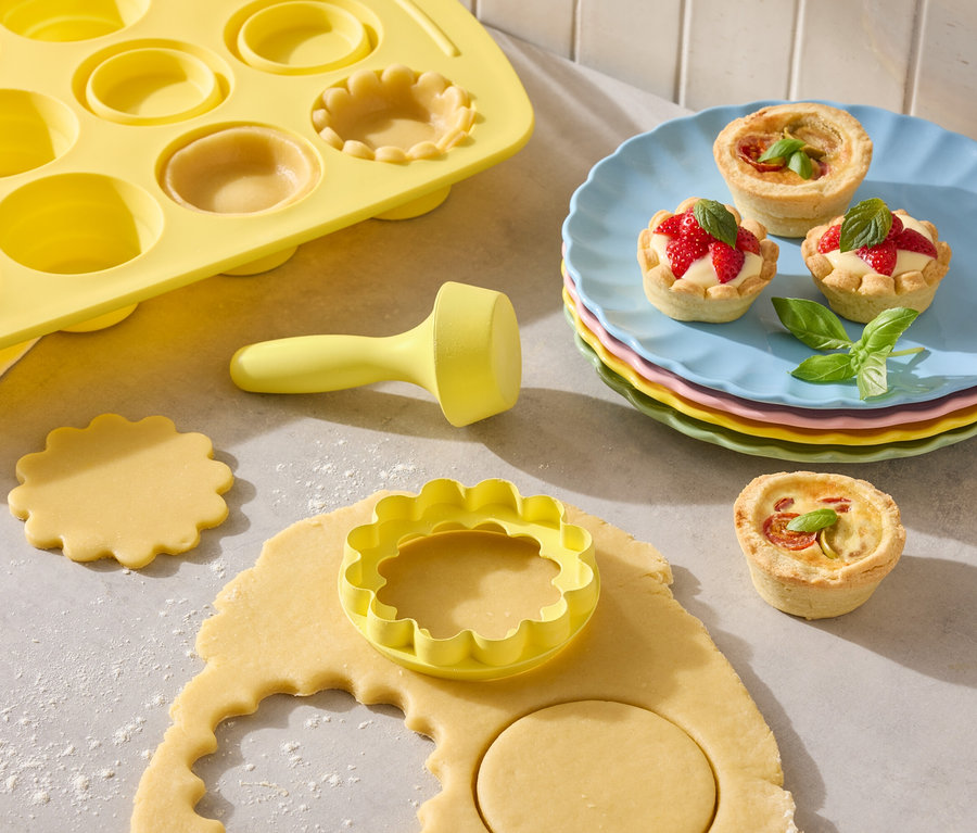 Gelbe Tarteform mit Ausstechern und fertige Tartelettes auf bunten Tellern.