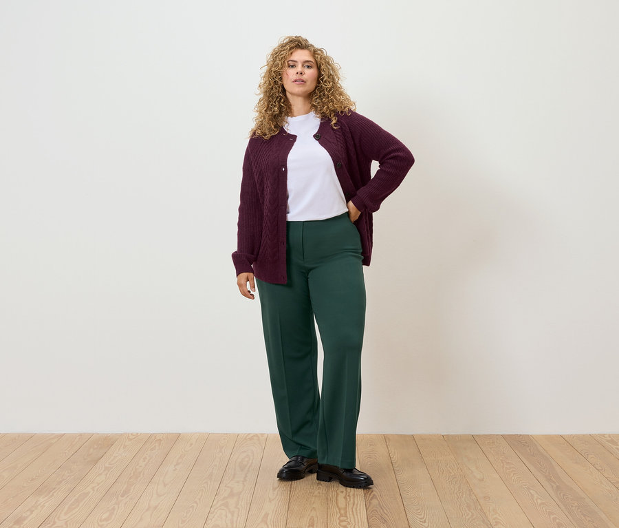 Eine Frau steht mit einer Hand in der Hüfte und trägt eine burgunderrote Strickjacke, ein weißes T-Shirt und eine grüne Hose.