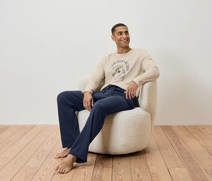 Ein Mann sitzt in einem Sessel und trägt ein beiges Langarmshirt und eine dunkelblaue Hose.