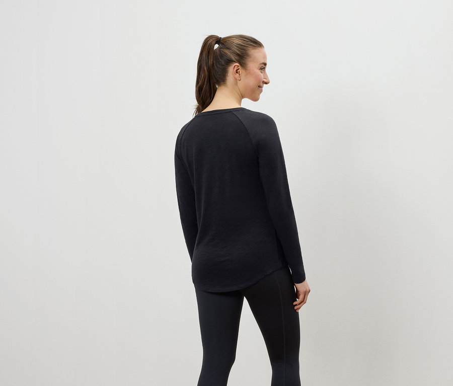 Frau mit braunen Haaren, die zu einem Pferdeschwanz zusammengebunden sind, posiert mit dem Rücken zur Kamera und trägt ein schwarzes Langarmshirt und schwarze Leggings.