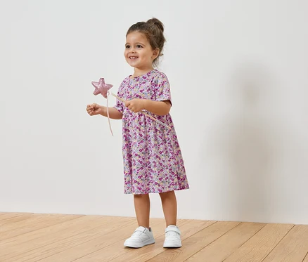 Lächelndes kleines Mädchen hält einen Sternenstab und trägt ein geblümtes Kleid und Turnschuhe.