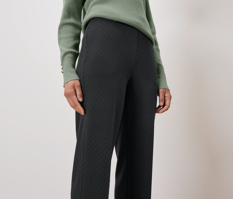 Eine Frau trägt einen grünen Pullover und eine schwarze, gemusterte Hose.