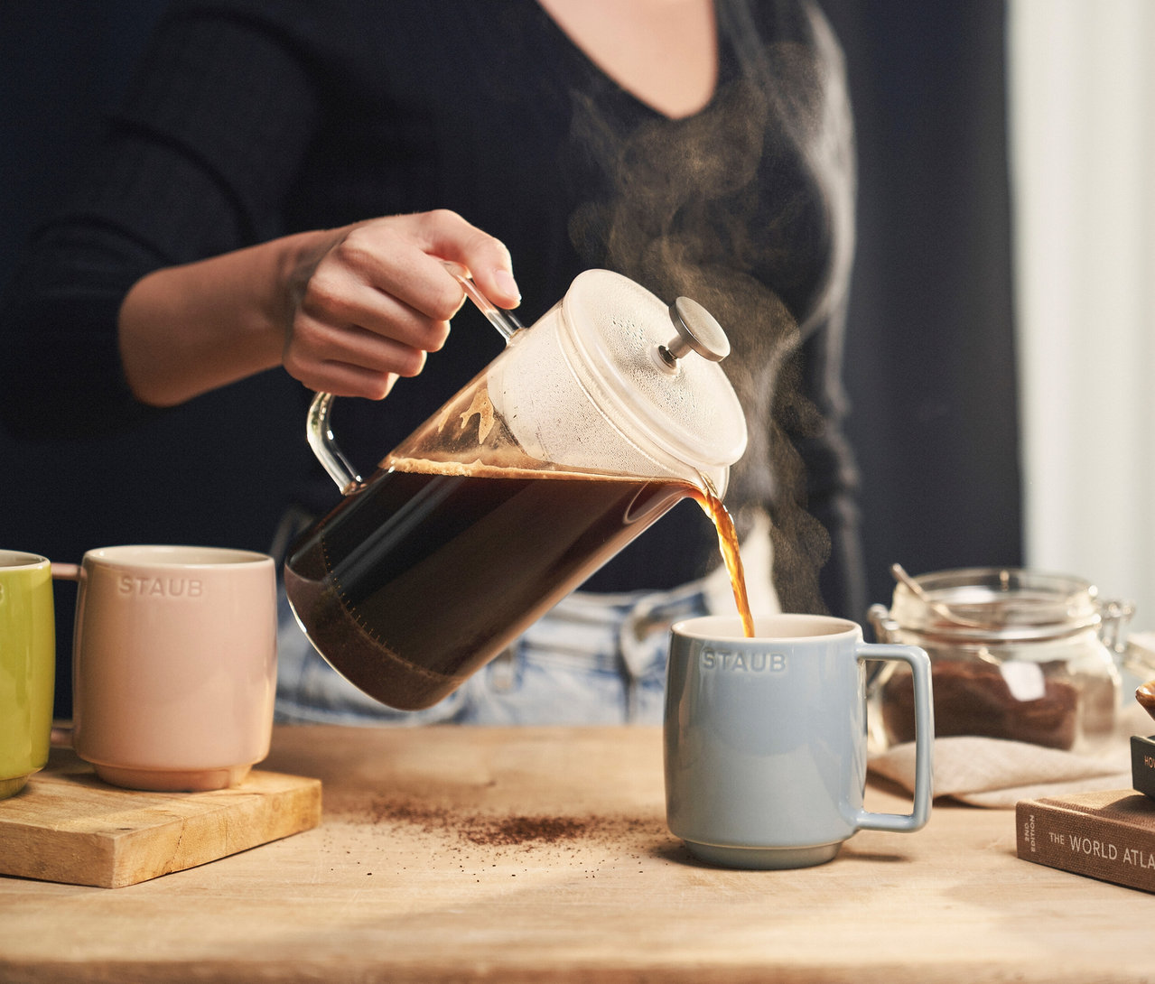 Frau gießt Kaffee aus einer Glaskanne in eine hellblaue STAUB Kaffeetasse. Daneben stehen weitere STAUB Kaffeebecher »Macaron« in Rosa und Grün.