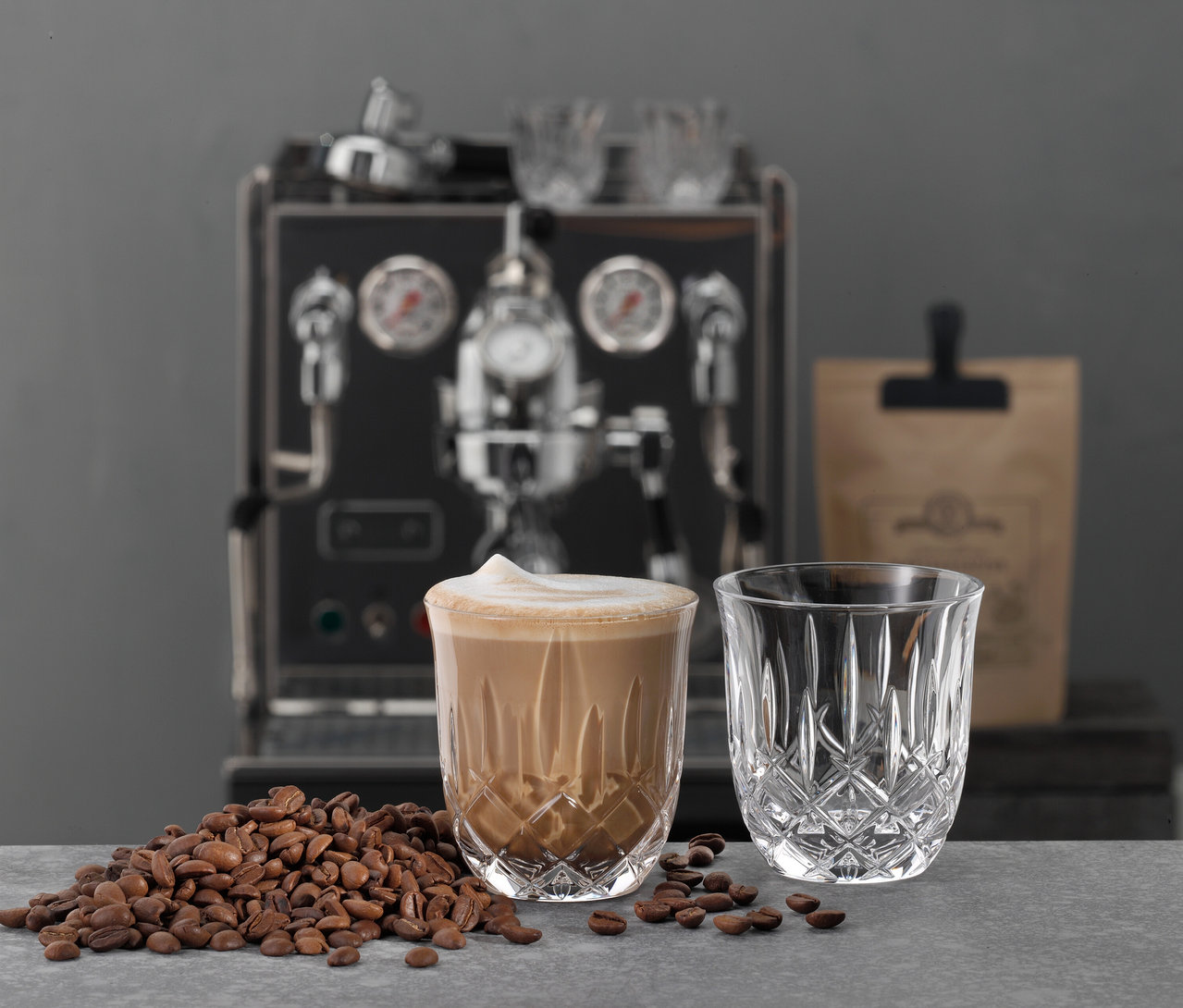 Ein mit Cappuccino gefülltes Nachtmann »Noblesse« Glas steht neben Kaffeebohnen. Im Hintergrund ist eine Kaffeemaschine.