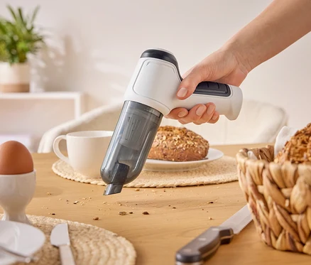 Eine Hand saugt Krümel mit einem weißen Handstaubsauger von einem Holztisch auf. Auf dem Tisch befinden sich auch ein Ei, eine Tasse und Brot.