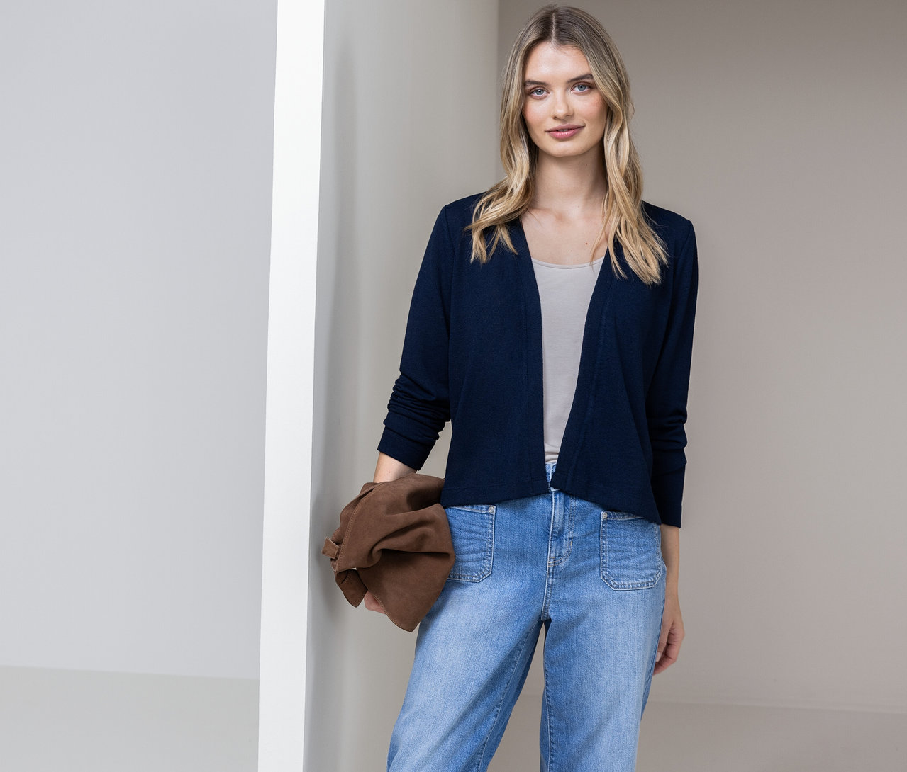 Frau mit blauem Street One Cardigan, Jeans und brauner Tasche vor einer Wand.