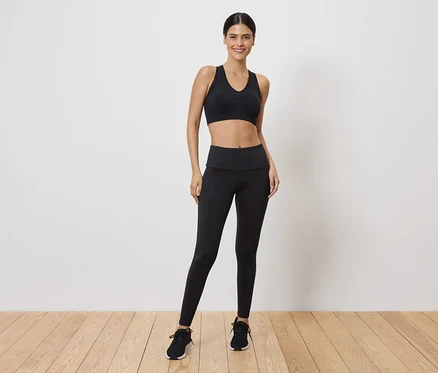 Frau in schwarzem Sport-BH und Leggings, stehend vor einer weißen Wand.