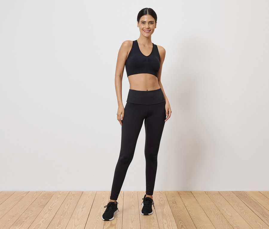 Frau in schwarzem Sport-BH und Leggings, stehend vor einer weißen Wand.