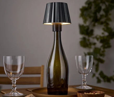 Eine Tischlampe in Form einer Weinflasche steht auf einem Holztisch neben zwei Weingläsern und einer Schale Nüsse.