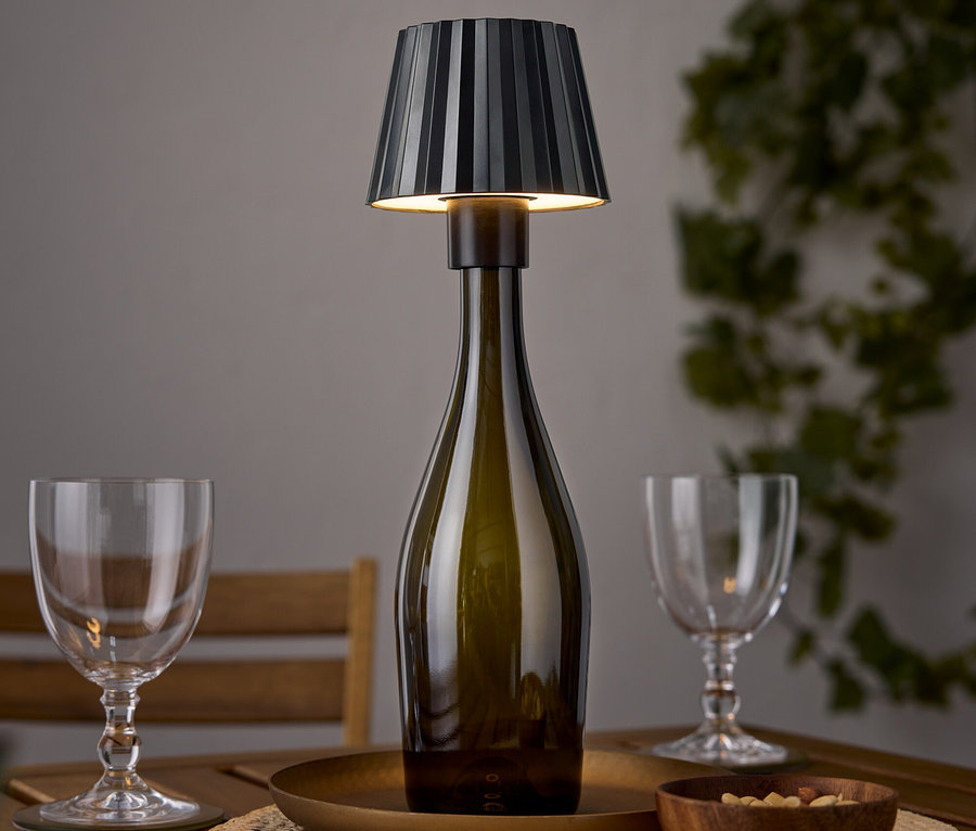 Eine Tischlampe in Form einer Weinflasche steht auf einem Holztisch neben zwei Weingläsern und einer Schale Nüsse.