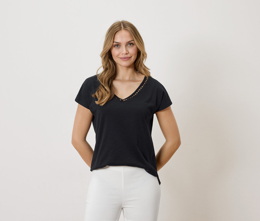 Frau mit blonden Haaren trägt ein schwarzes T-Shirt mit V-Ausschnitt und weißen Hosen vor einem neutralen Hintergrund.