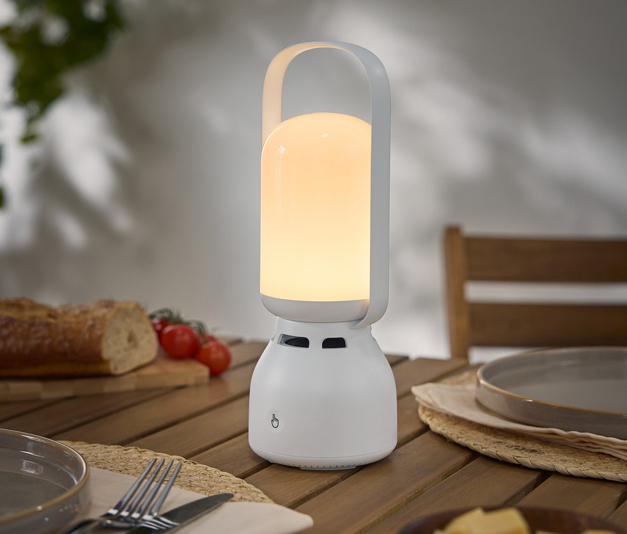 Eine eingeschaltete, tragbare, weiße Lampe mit Henkel steht auf einem Holztisch neben einem Teller, Besteck, Brot, Tomaten und Käse.