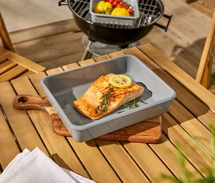 Ein Stück Lachs liegt in einer grauen Auflaufform auf einem Holztisch, daneben ein Grill.