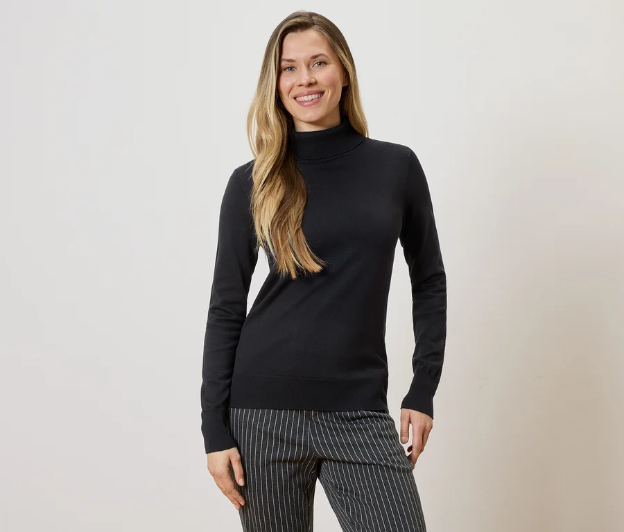 Frau mit langen blonden Haaren, die einen schwarzen Rollkragenpullover und eine gestreifte Hose vor einem hellen Hintergrund trägt.