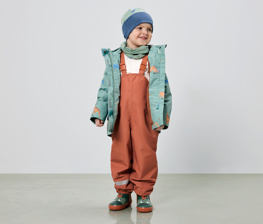 Ein lächelnder Junge trägt eine grüne Jacke mit Dinosaurier-Muster, eine orangefarbene Latzhose, einen blauen Hut und grüne Gummistiefel.