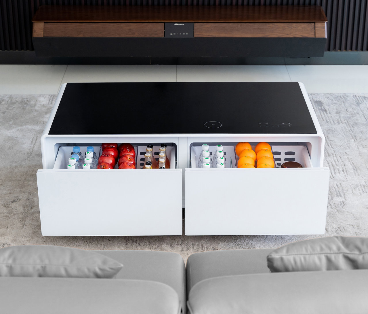 Ein weißer Caso Sound & Cool Tisch mit Getränken und Obst in offenen Schubladen steht vor einem grauen Sofa.
