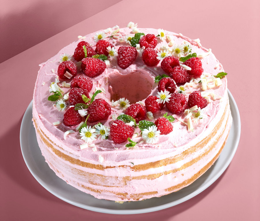 Mehrschichtiger Kuchen mit rosa Creme, Himbeeren, Blüten und Spänen auf einem weißen Teller.