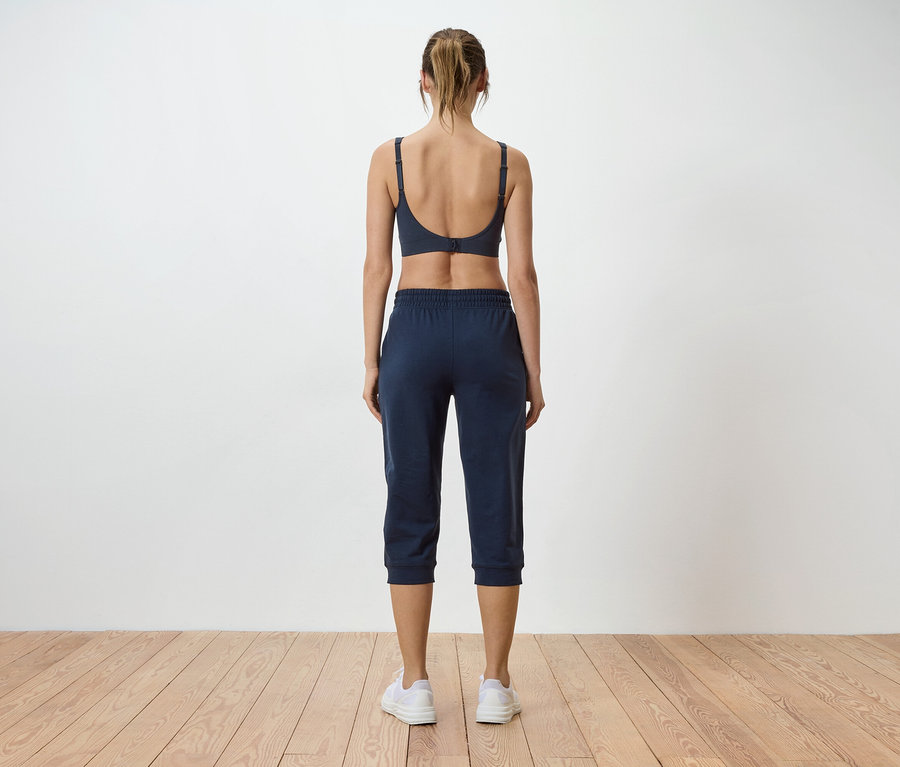 Rückansicht einer Frau mit zusammengebundenen blonden Haaren, die einen grauen Sport-BH und eine blaue Dreiviertelhose vor einer weißen Wand trägt.