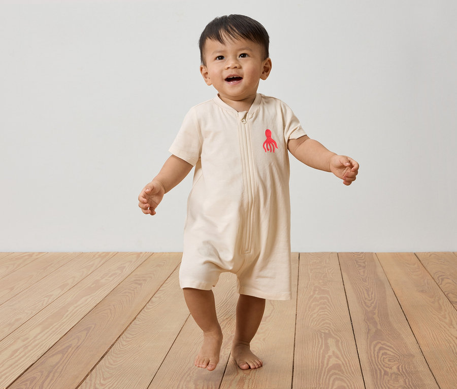 Ein Kleinkind geht barfuß auf einem Holzboden und trägt einen beigen Overall mit einem roten Tintenfisch darauf.