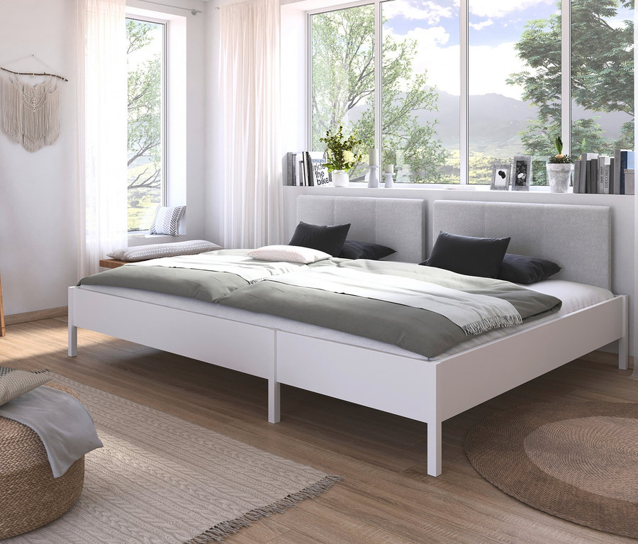 Weißes Doppelbett mit grauer Bettwäsche, schwarzen Kissen und zwei Kopfteilen vor einem Fenster mit Blick auf Bäume.