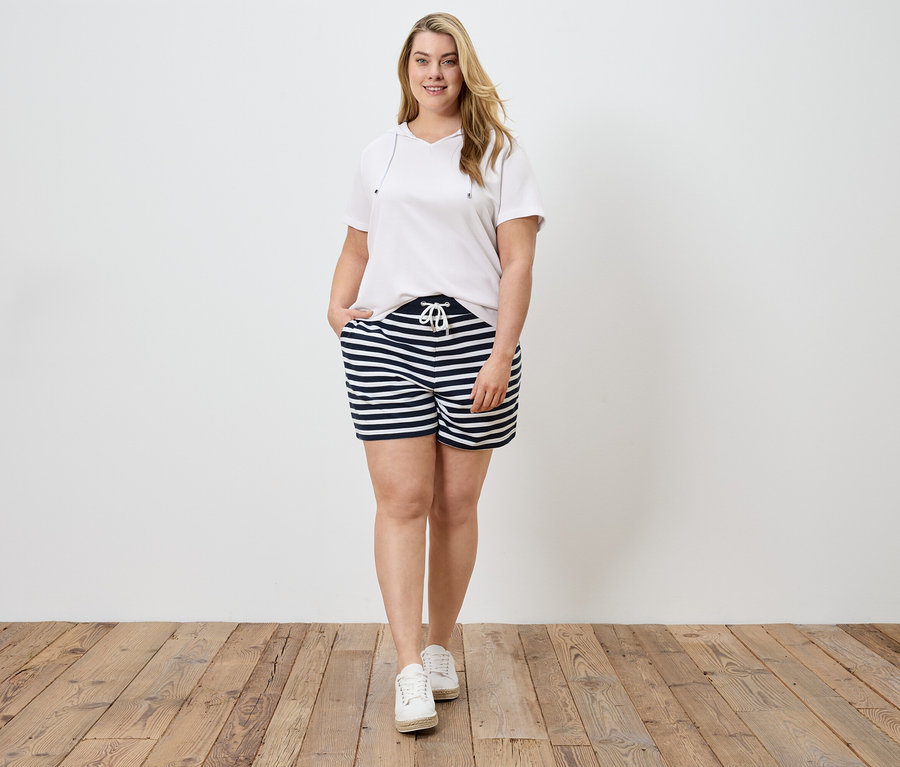 Frau in weißem T-Shirt und gestreiften Shorts mit Hand in der Tasche, vor weißer Wand und Holzboden.
