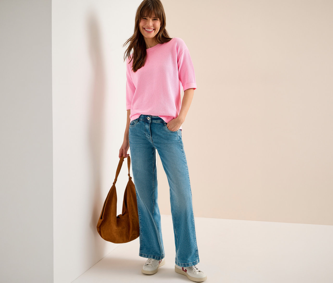 Frau mit brauner Tasche, blauen Jeans und CECIL Kurzarmpullover in Pink. Sie steht vor einer Wand.