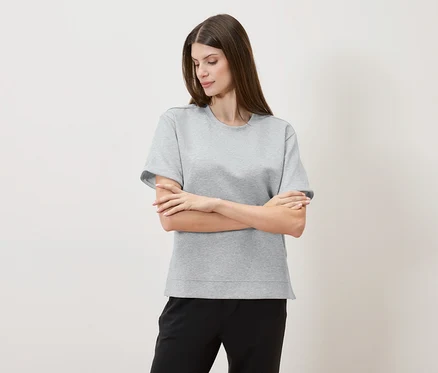 Frau mit verschränkten Armen trägt ein graues T-Shirt und eine schwarze Hose vor einem neutralen Hintergrund.