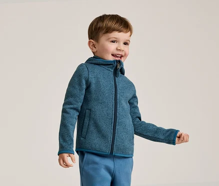 Lächelnder Junge mit braunen Haaren trägt eine blaue Fleecejacke mit Kapuze und blaue Jogginghose vor einem hellen Hintergrund.