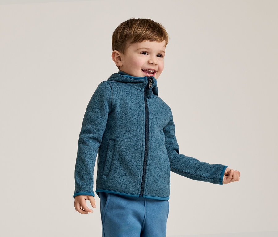 Lächelnder Junge mit braunen Haaren trägt eine blaue Fleecejacke mit Kapuze und blaue Jogginghose vor einem hellen Hintergrund.