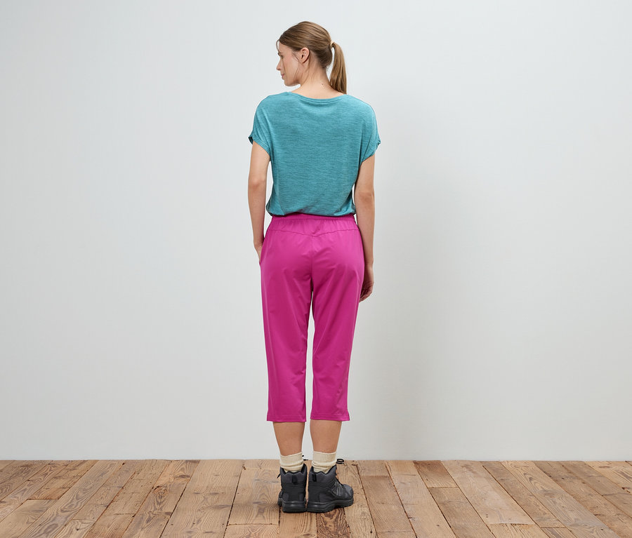 Eine Frau steht mit dem Rücken zur Kamera, sie trägt ein türkisfarbenes T-Shirt, eine pinke Hose und dunkle Schuhe.