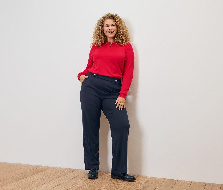 Frau mit lockigem Haar, rotem Oberteil und blauen Hosen.